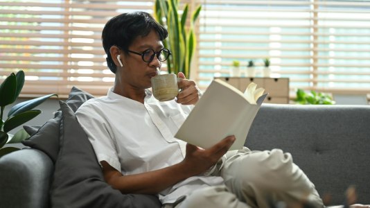 Peaceful asian man drinking hot coffee and reading book on comfortable couch.